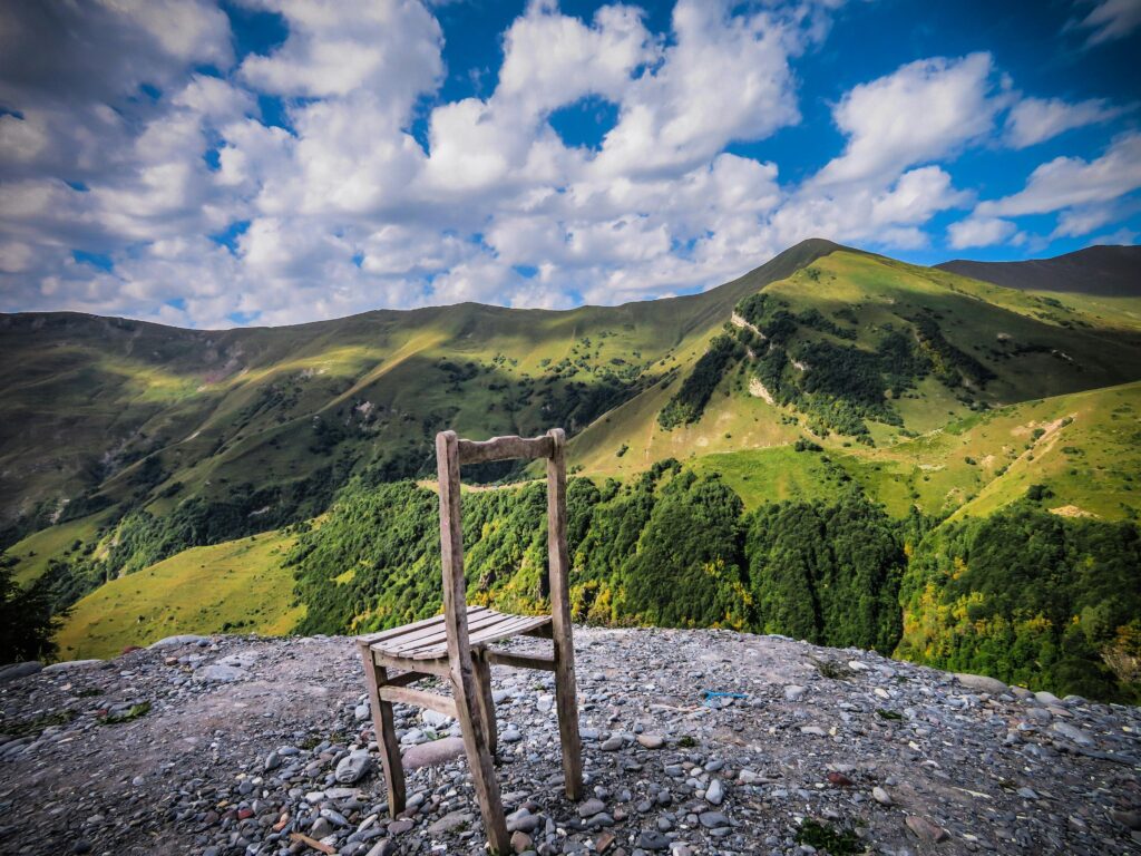 A wooden chair placed on a hillside with a panoramic view of lush Georgian mountains under a vibrant sky.